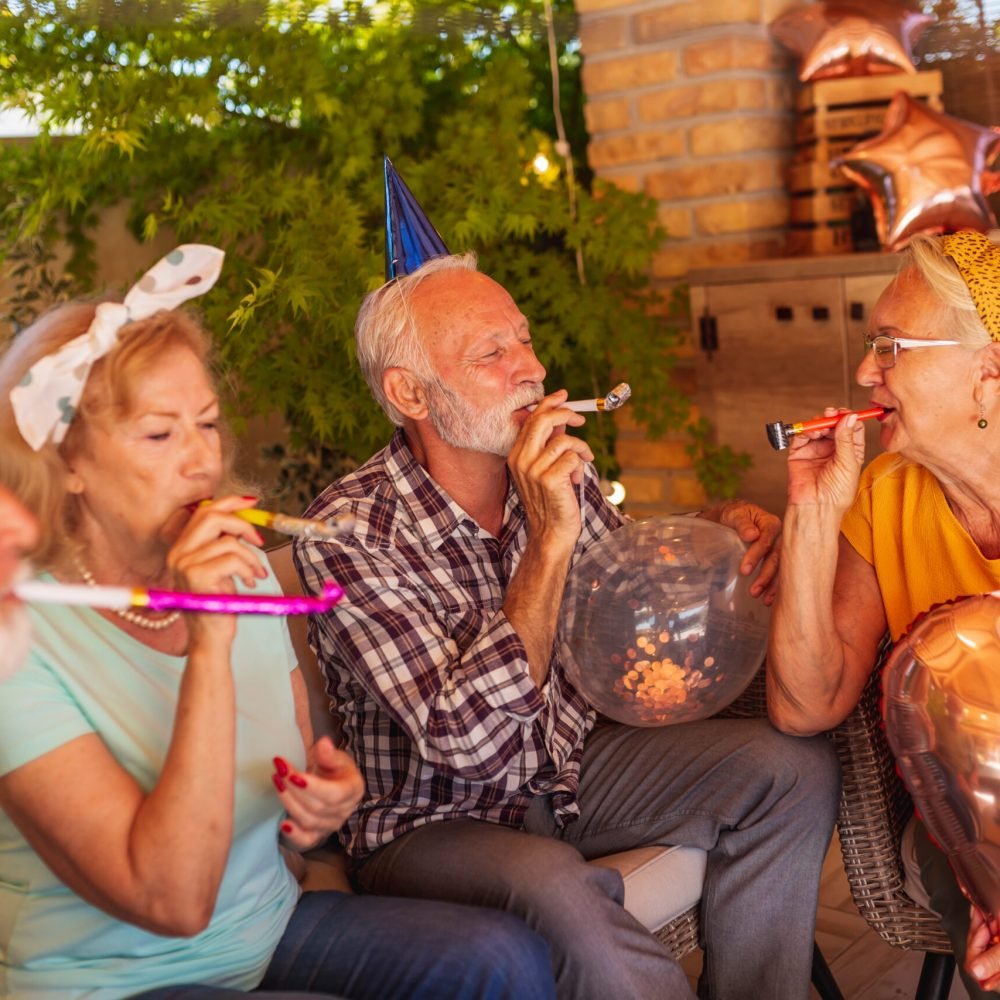 Group of cheerful elderly people having fun at a birthday party, holding balloons and blowing party whistles