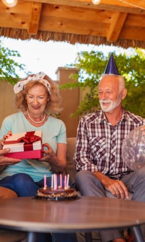Group of cheerful elderly people having fun at a birthday party, wearing party hats, holding balloons and opening presents