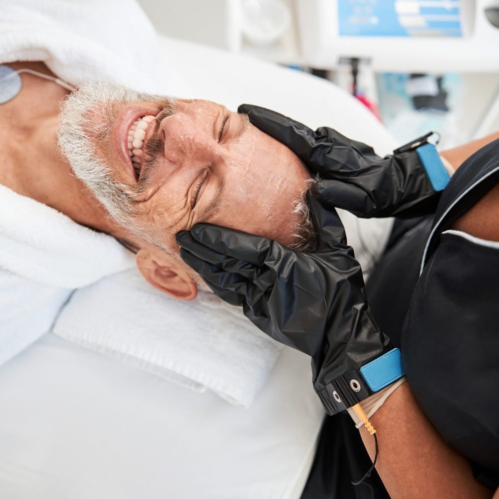 Cheerful elder man smiling while laying on a spa table and having a facial massage